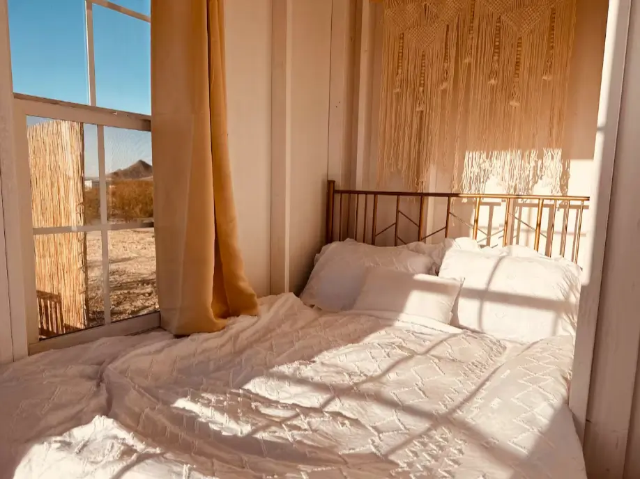 A cozy bed in a one bedroom tiny house, featuring soft white bedding and a macrame wall hanging, creating a peaceful sanctuary.