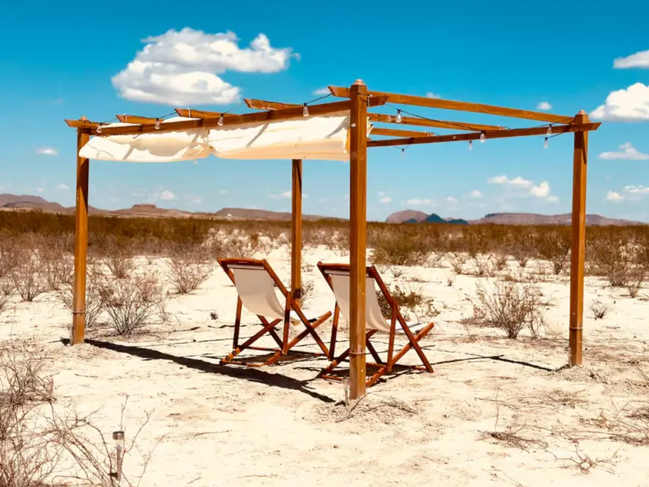 A simple wooden pergola with lounge chairs creates an outdoor living room for a one bedroom tiny house in the desert.