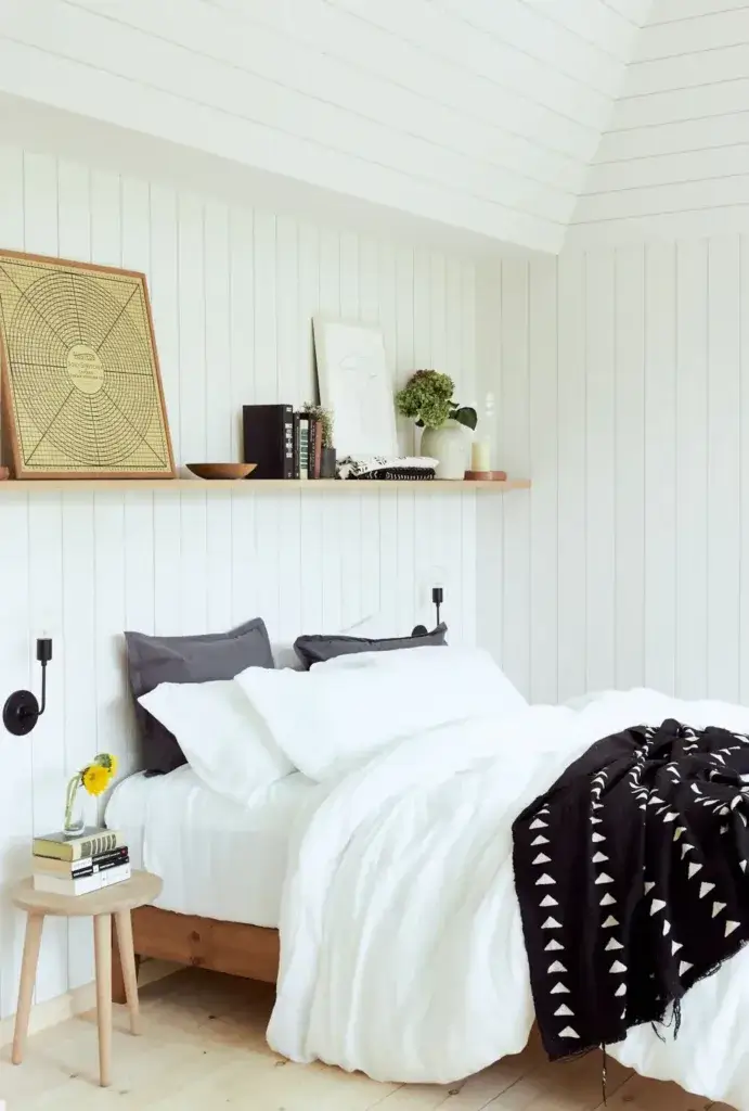 A cozy bedroom in a small footprint home using a floating shelf as a headboard and wall-mounted lights to save space.
