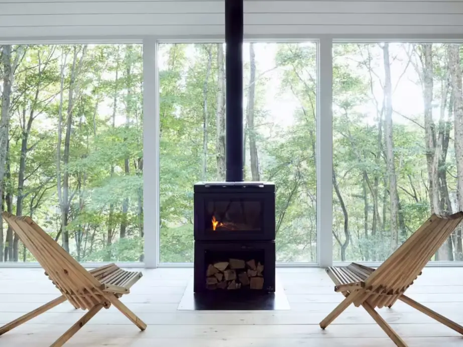 A modern wood-burning stove in front of large windows looking out into a forest, symbolizing the cozy and simple lifestyle of tiny home living.