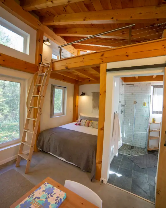 A view of the main floor bedroom in the Honeycrisp Cottage, showing the loft above, a wooden ladder for access, and a sliding barn door opening to the modern tiny house bathroom.