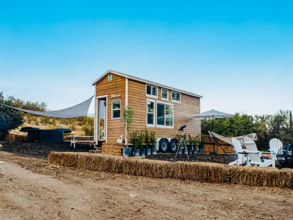 The outdoor living space of a tiny house, featuring a picnic table, adirondack chairs, and hay bales to extend the home's functional area.