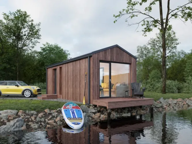 The Nordic Mini modular cabin with wood siding on a waterside deck, a Scandinavian prefab home under 50k.