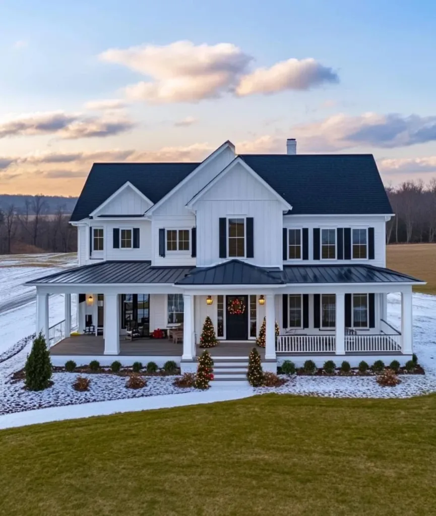 White farmhouse with a wrap-around porch decorated for Christmas with wreaths and small trees.