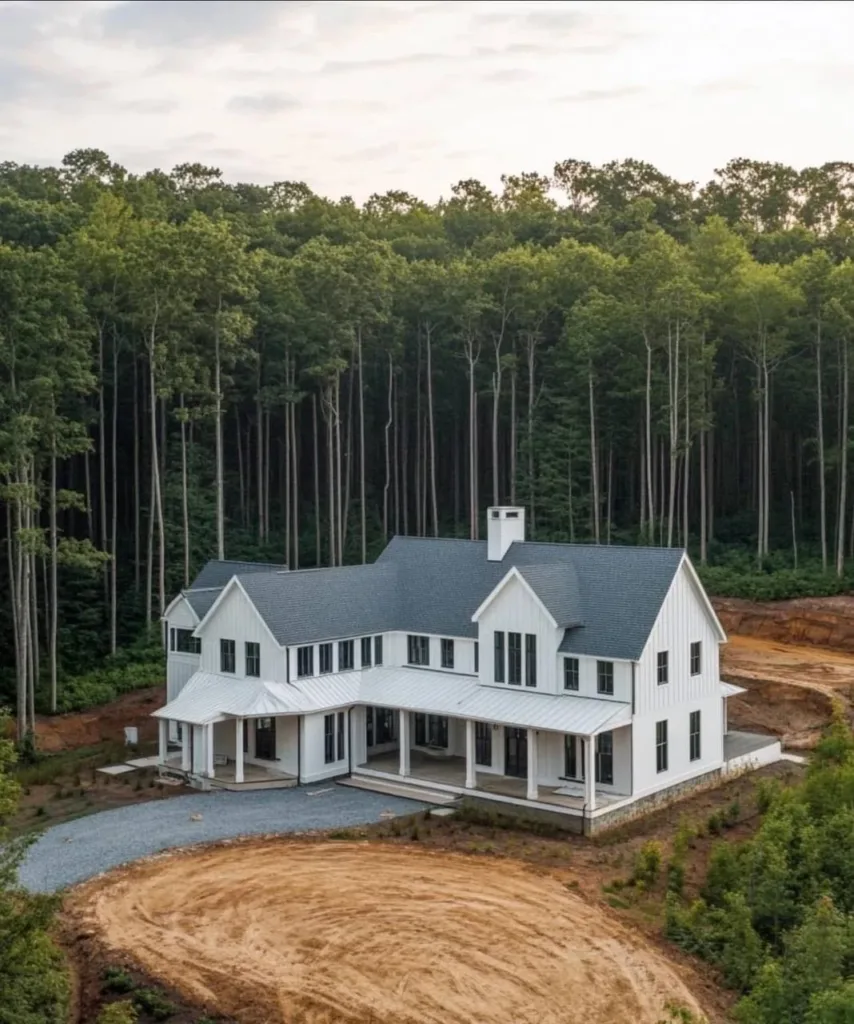 White farmhouse with a gray roof and wrap-around porch, nestled among tall trees.