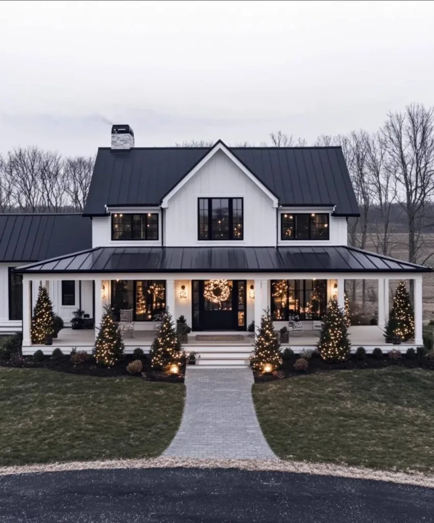 White farmhouse with black trim, a wrap-around porch decorated with miniature Christmas trees, and a wreath on the door.