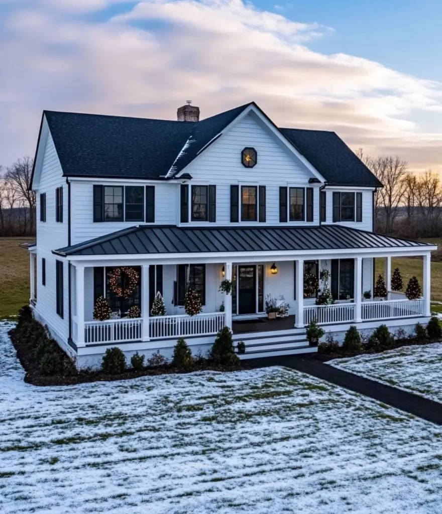 Modern farmhouse with white siding, black trim and awning, a wrap-around porch decorated for Christmas, and a light dusting of snow on the ground.