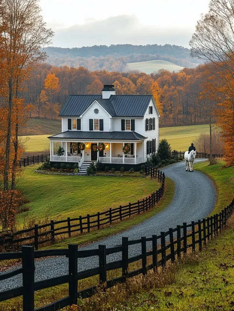 Modern farmhouse with white siding, a black roof, and a wrap-around porch, with a winding gravel driveway, a wooden fence, and fall foliage in the background.