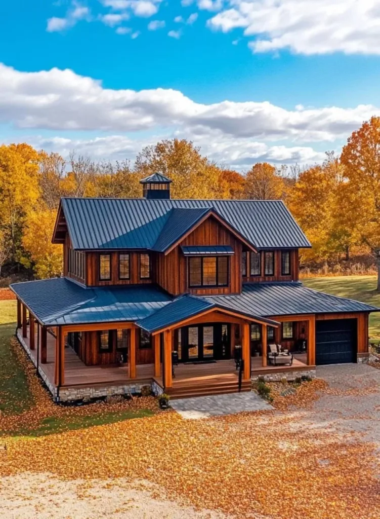 Barndominium with natural wood siding, a blue metal roof, a cupola, a wrap-around porch, and a stone base, surrounded by fall foliage.