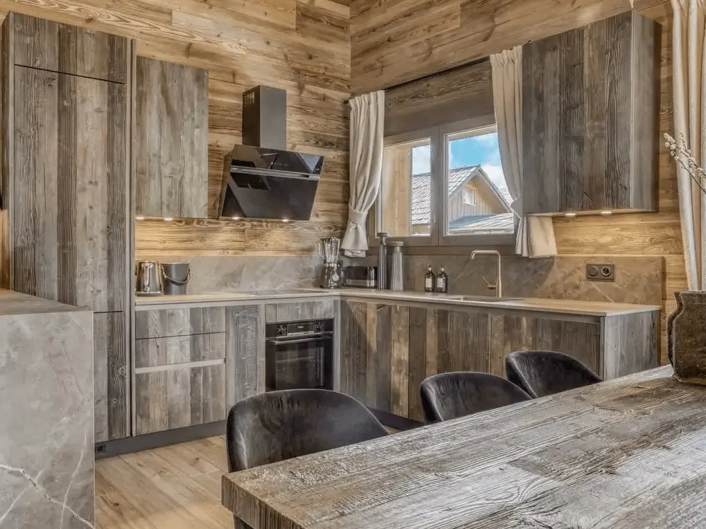 A modern rustic cabin interior with weathered wood kitchen cabinets, gray countertops, and a large dining table in the foreground.