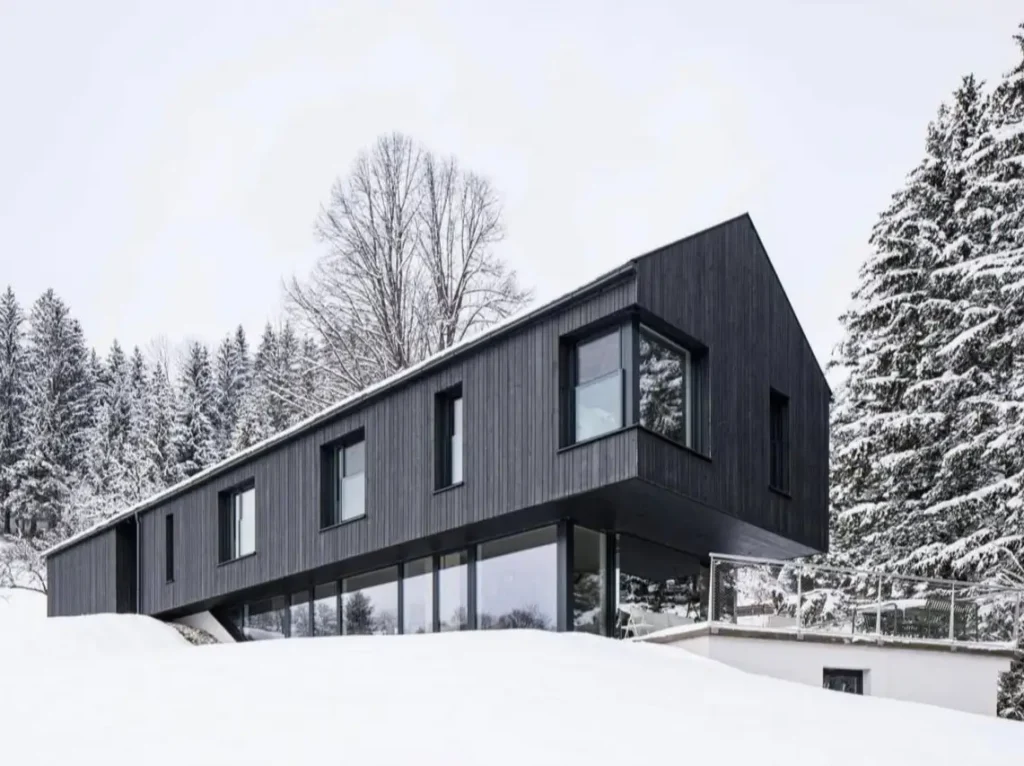 A modern minimalist luxury mountain house with black siding and large windows in a snowy forest.