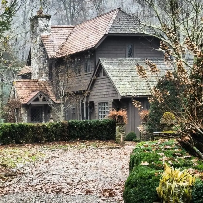 A rustic cottage-style mountain house with dark siding and a stone chimney, surrounded by trees in the mist.
