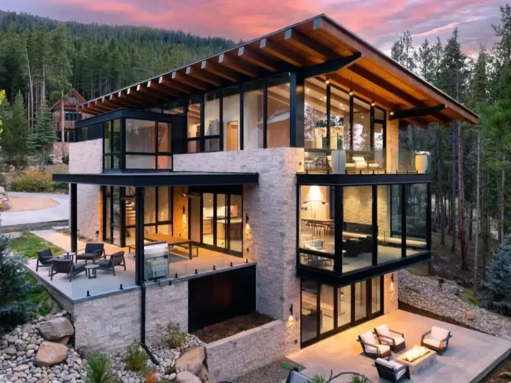 A luxury modern mountain home with stone walls, glass exteriors, and multiple patios at sunset.