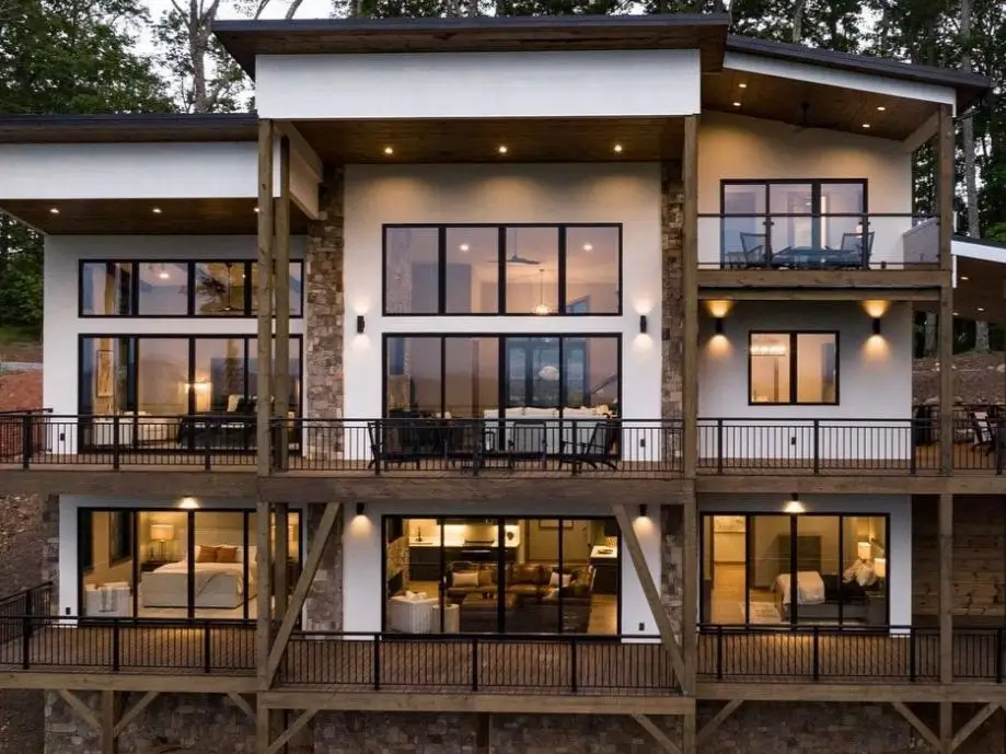 The back exterior of a multi-story modern mountain home with three levels of glowing decks at dusk.