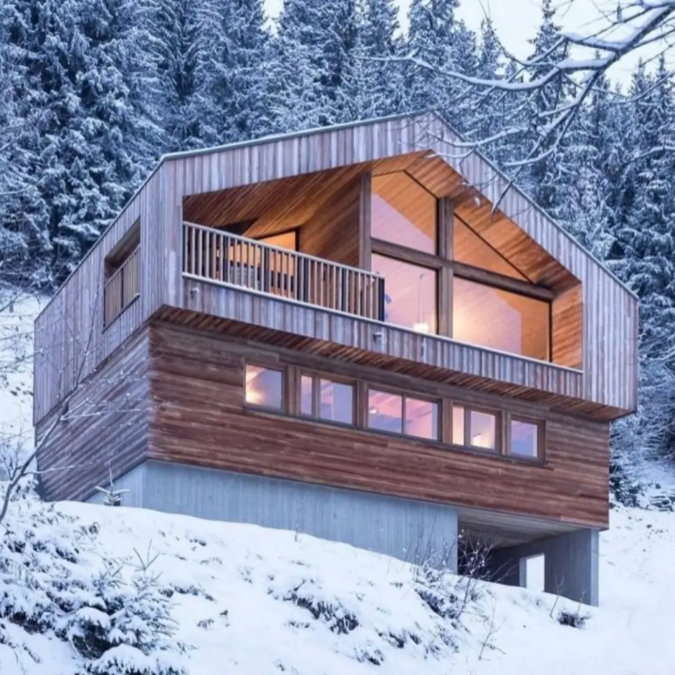 A modern wood-clad mountain home with an inset balcony, set in a snowy forest landscape.