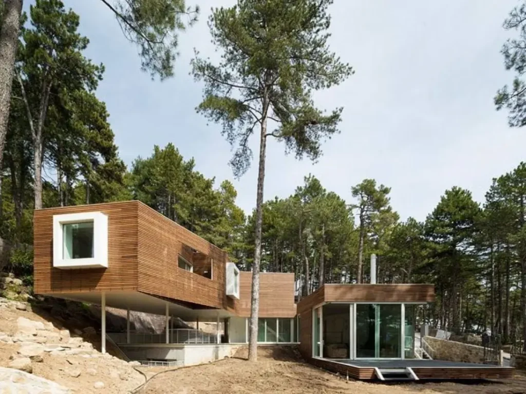 A modern wood-clad mountain home with a cantilevered upper level on stilts in a forest.