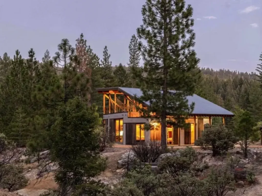 A modern rustic mountain cabin with an exposed wood frame, glowing from within at dusk in a pine forest.