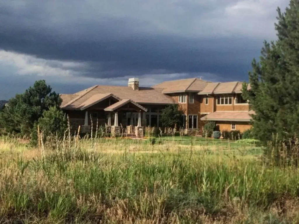 A large ranch-style mountain home surrounded by tall prairie grass under a dark, stormy sky.