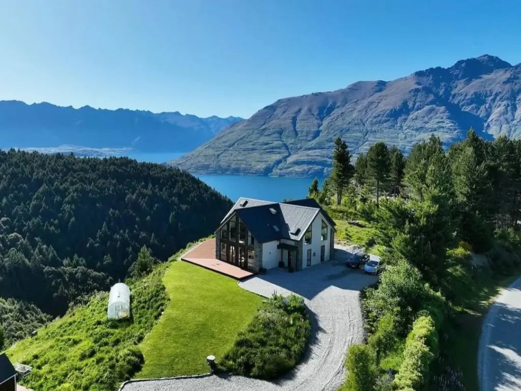 An aerial view of a modern luxury mountain home with a deck overlooking a vast lake and mountain range.