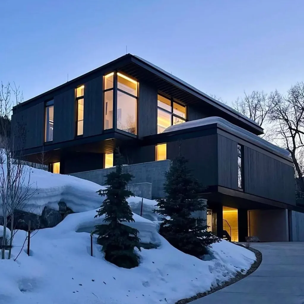 A contemporary luxury mountain home with dark siding and glowing windows at dusk in the snow.