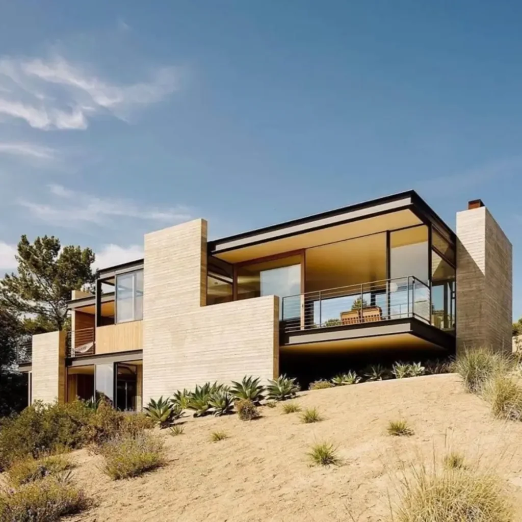 A modern desert mountain home with light brick, glass walls, and a balcony on a sandy hill.