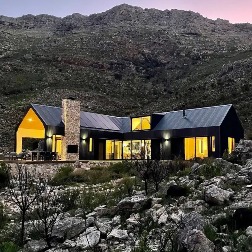 A modern black barn-style mountain home with a large stone chimney, nestled in a rocky landscape at twilight.
