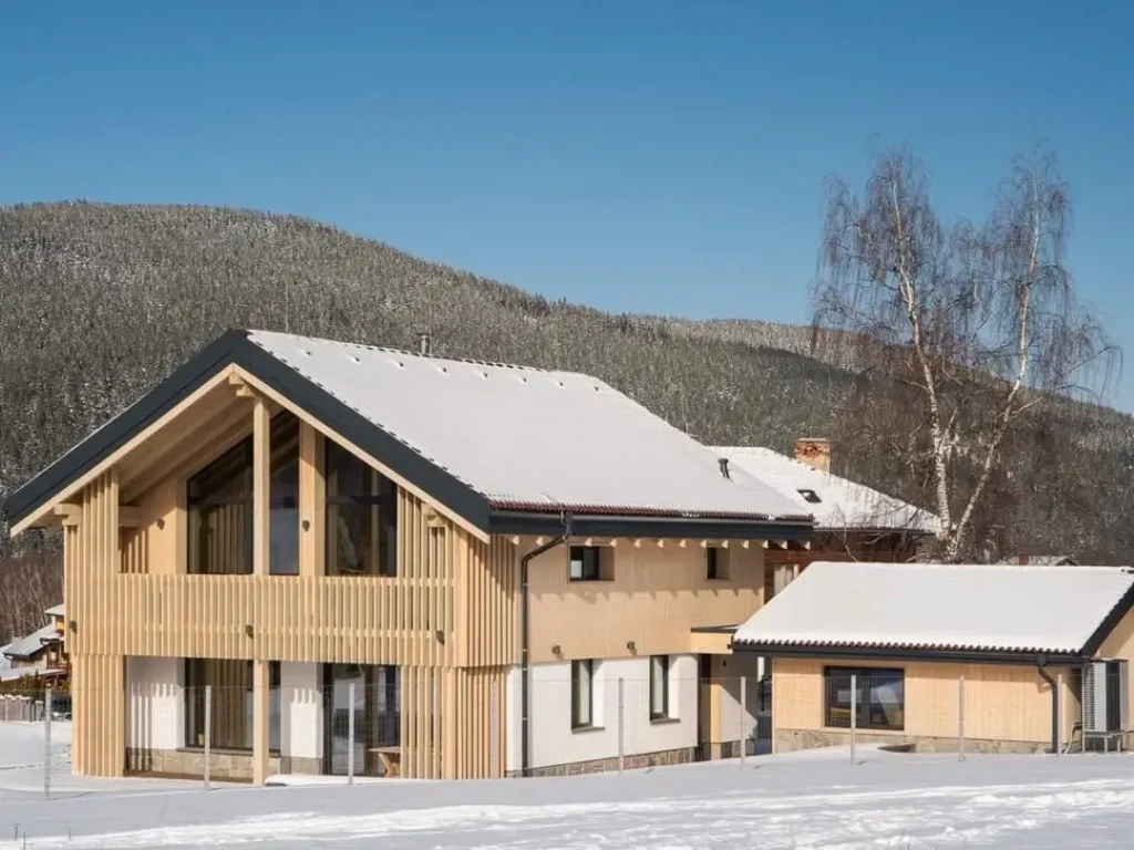 A modern mountain home with light vertical wood siding and a dark roof in a snowy setting.