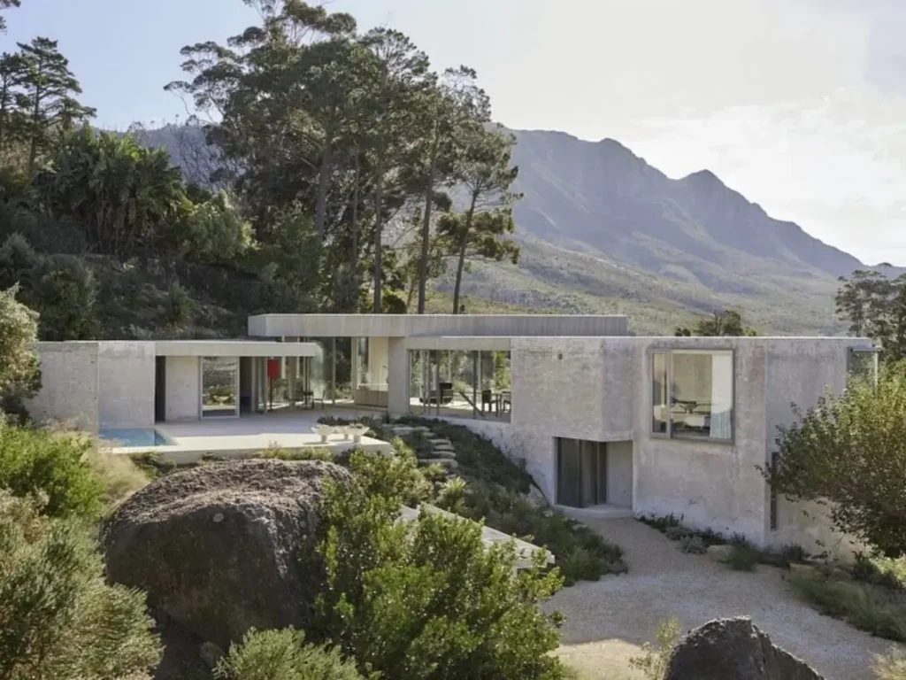 A modern concrete minimalist house with large windows, set among boulders with mountains in the background.