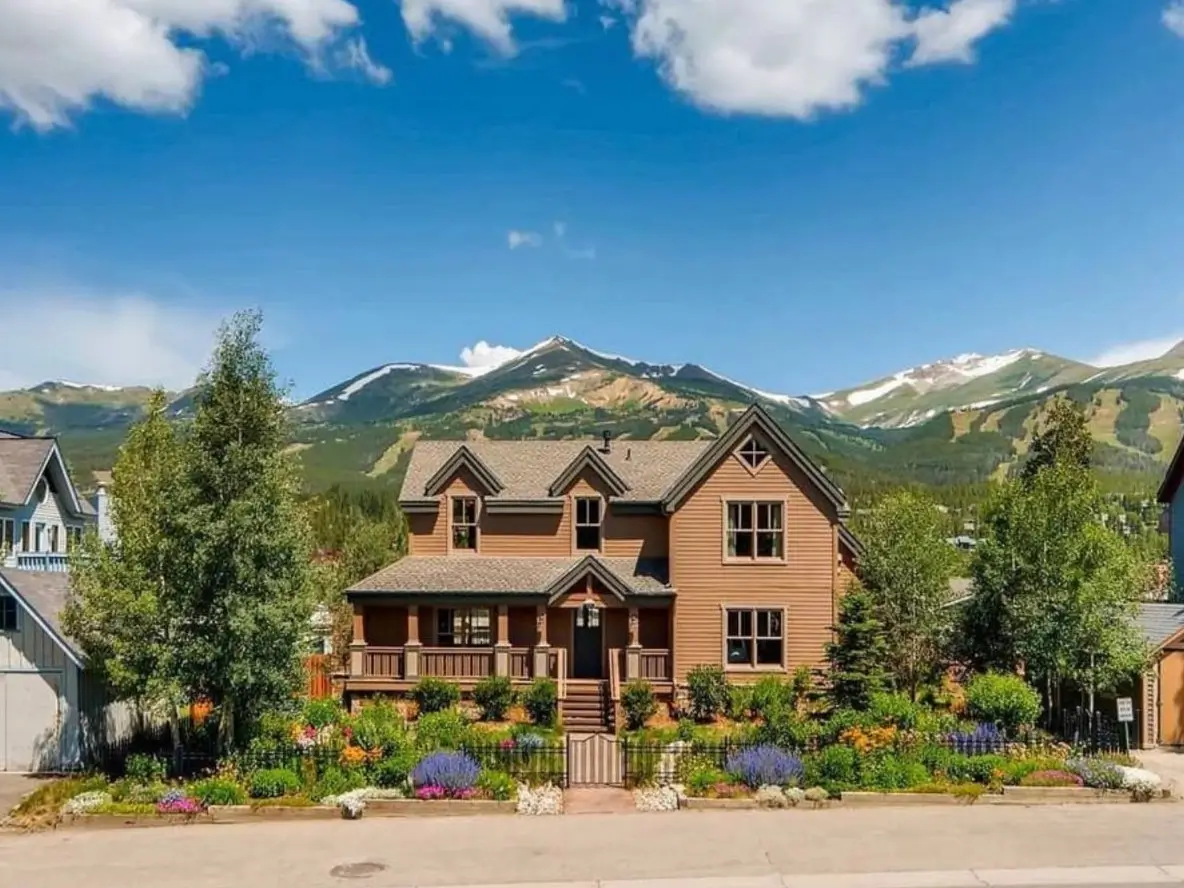 Exterior of a classic luxury mountain house with a vibrant flower garden, set against a panoramic view of snow-capped mountains under a blue sky.