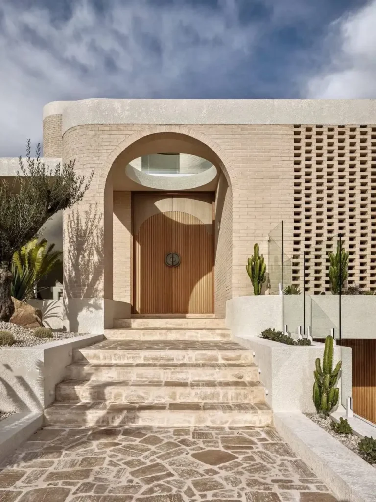 Detailed view of modern Mediterranean entrance showing arched wood door, textured light brick, stone paving, and architectural screen.