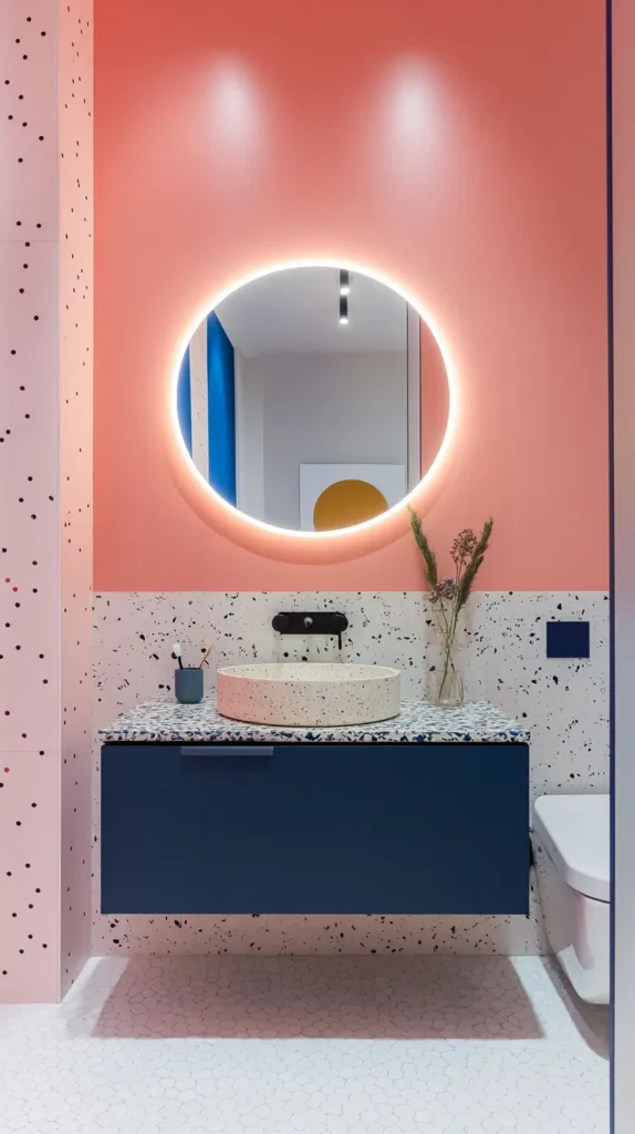 Playful modern bathroom vanity area featuring a dark blue floating cabinet, a white terrazzo countertop and backsplash with large dark flecks, a round speckled vessel sink, a round backlit mirror, against a peachy-pink painted upper wall.