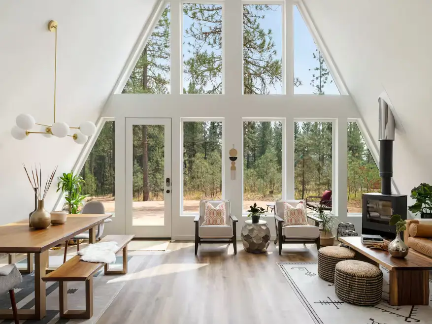 Sun-drenched modern A-frame living space with a full wall of triangular windows, light wood floors, white walls, and minimalist contemporary furniture.