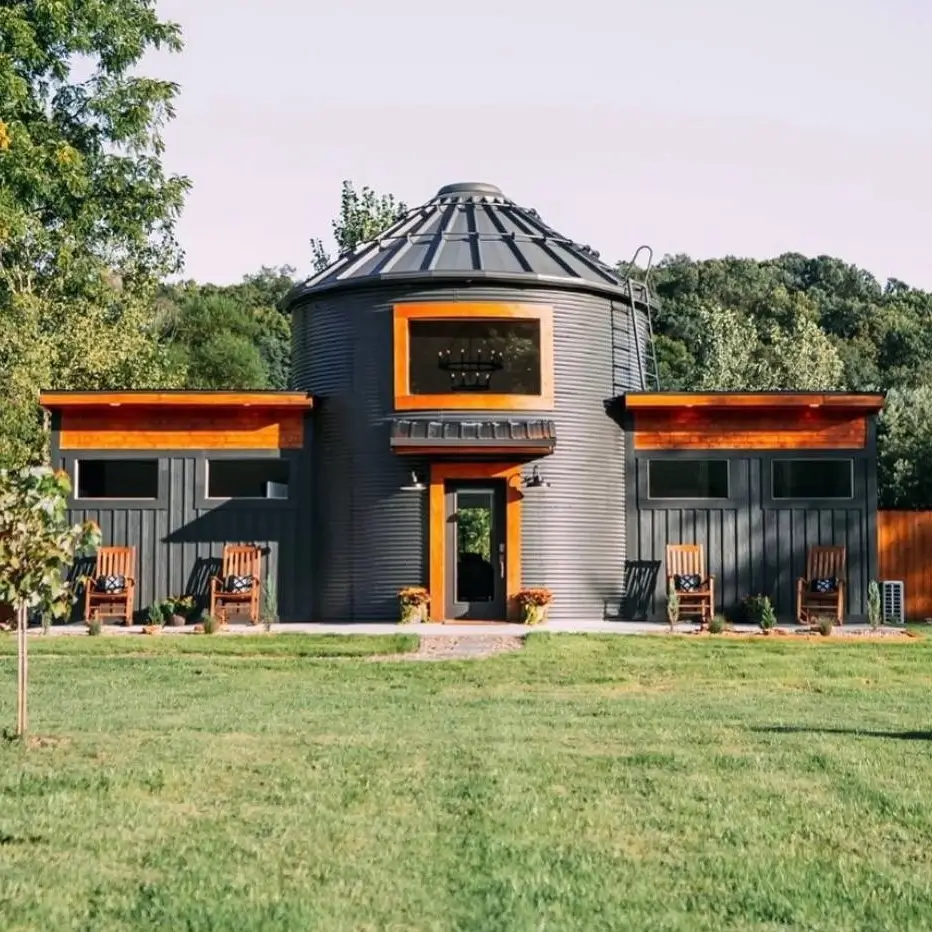 A dark grey, two-story grain silo house stands as the central point between two symmetrical, single-story wings with wood accents and large windows.