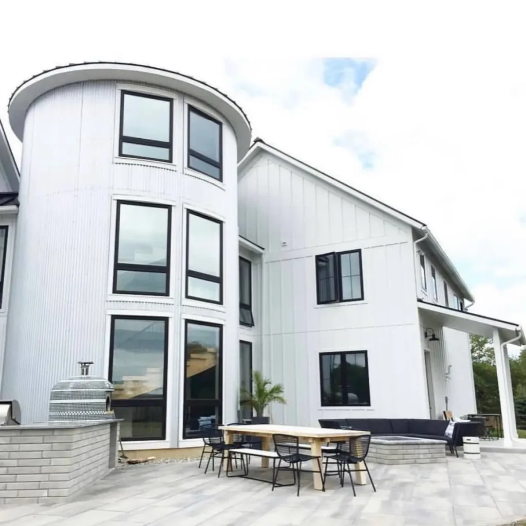 A large, multi-story modern farmhouse painted in crisp white features an integrated silo-shaped tower with large, black-framed windows, connected to a spacious outdoor patio.