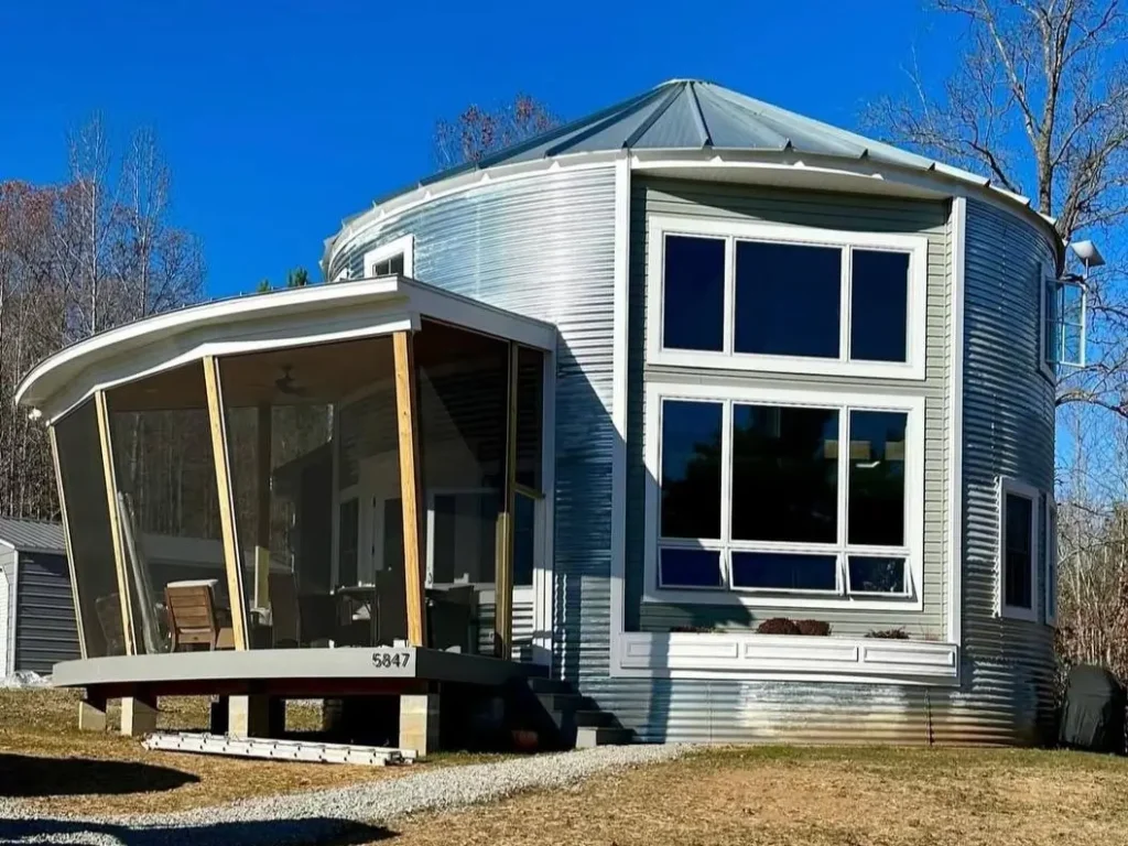 A classic, unpainted galvanized steel grain silo home features large residential windows and a gracefully curved, screened-in porch addition.