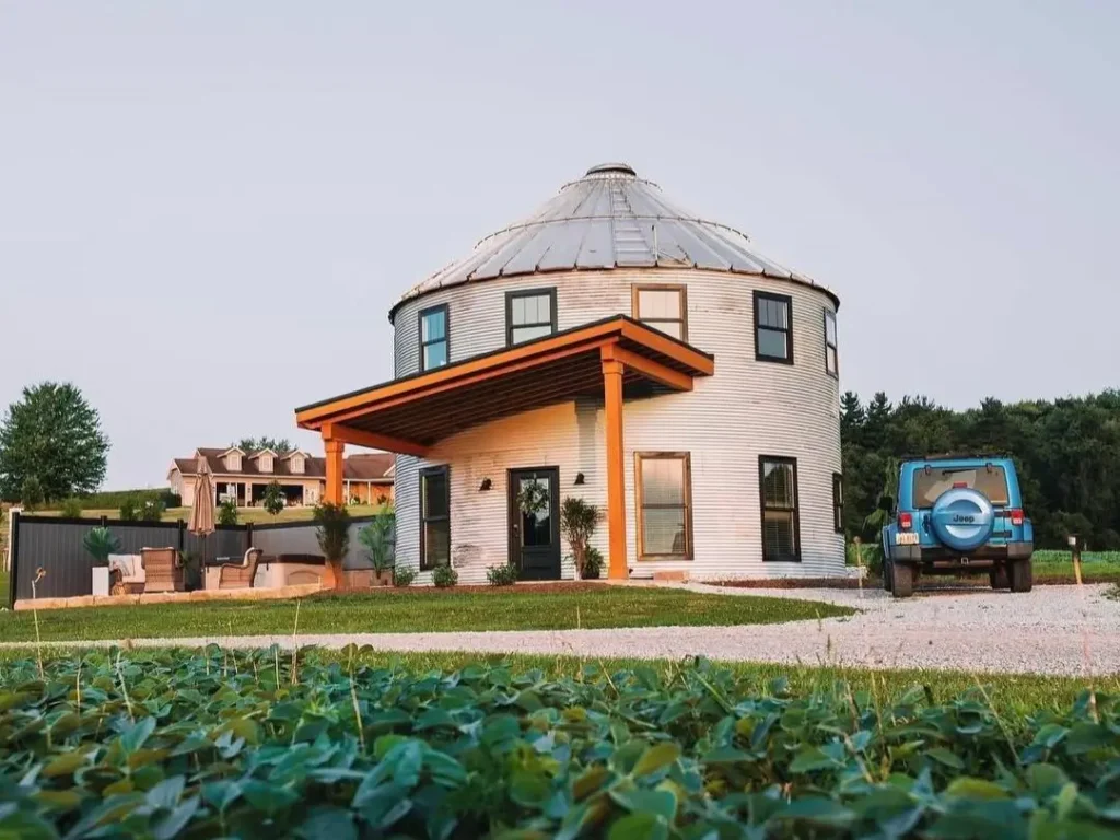 A quaint, white grain silo house with a simple wooden porch sits in a lush green field, evoking a sense of peaceful, rural living.