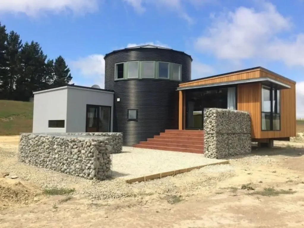 A modern home design features a central black grain silo connected to a wood-clad structure and a grey stucco wing, with rugged gabion walls defining the entryway.