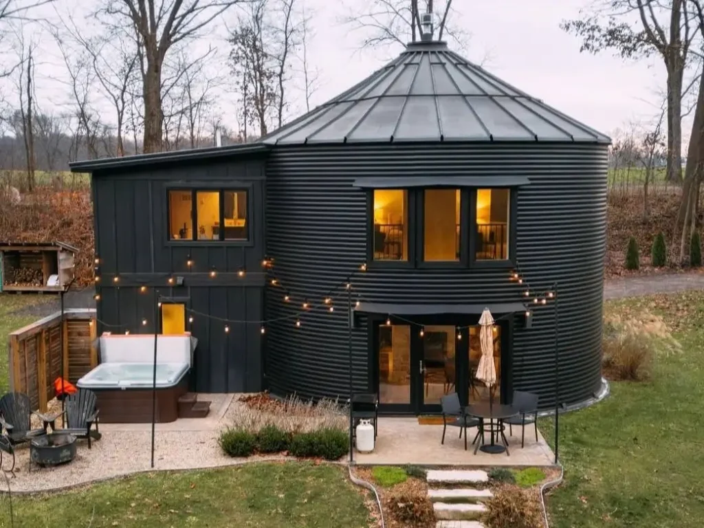 A dark, modern grain silo house at dusk, with warm lights glowing from the windows, an attached rectangular addition, and a cozy patio with a hot tub and string lights.