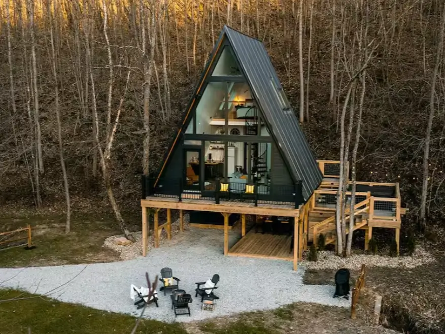 An elevated black A-frame treehouse in a forest setting, showcasing its multi-story interior and lower fire pit area.