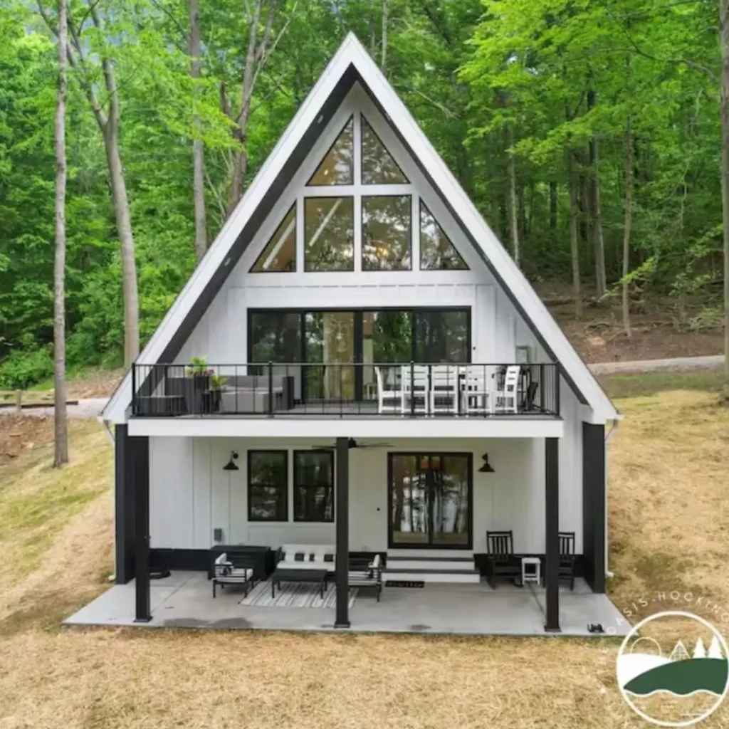 A modern two-story white A-frame cabin in Ohio with a black trim, an upper balcony, and a lower patio.