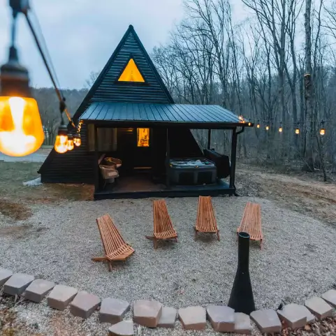 A secluded black A-frame cabin at dusk with a stone fire pit area and glowing string lights.