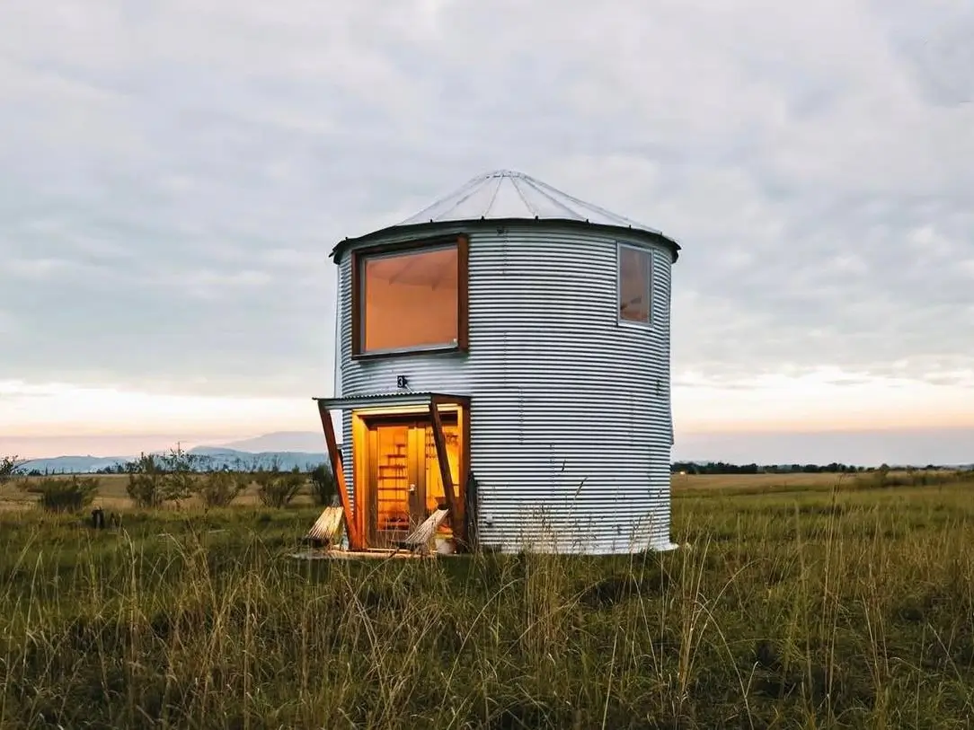 A modern grain silo house stands alone in a grassy field at dusk, with warm light glowing from its large windows and entryway.