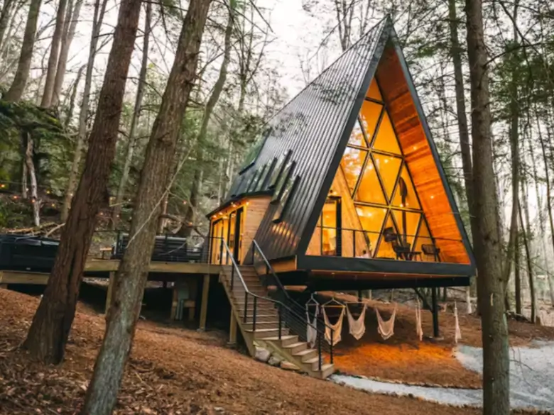 A luxury modern elevated A-frame cabin in Hocking Hills, glowing warmly at dusk with a full glass front and hammocks hanging underneath.